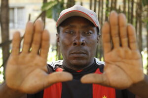 Foto colorida de homem negro, usa boné e camiseta de time com listras em preto e vermelho, suas mãos estão estendidas com a palma virada para frente.