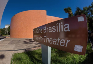 foto colorida em ambiente externo de uma placa marrom escrito cine brasília e o cinema atrás.